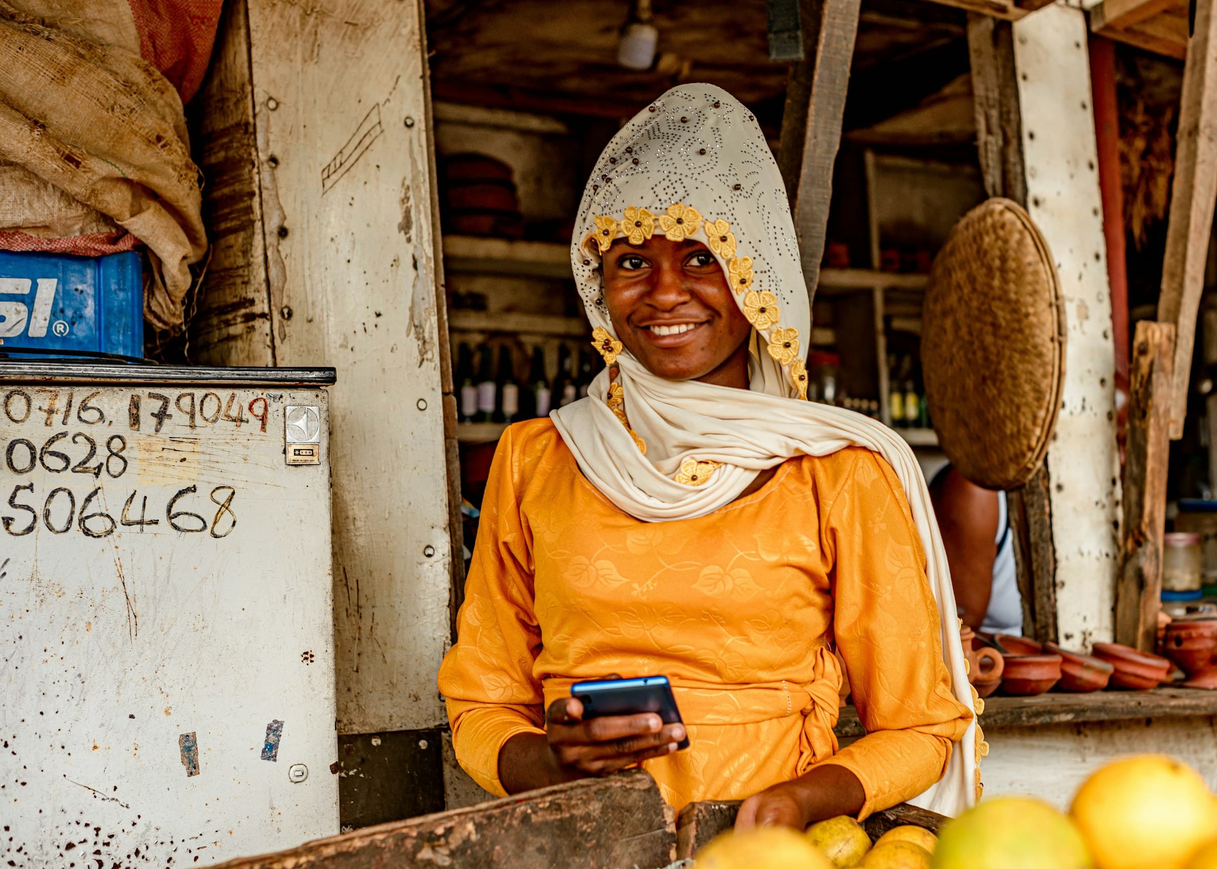 A shopkeeper in Dar es Salaam smiling and holding her phone at her market stall